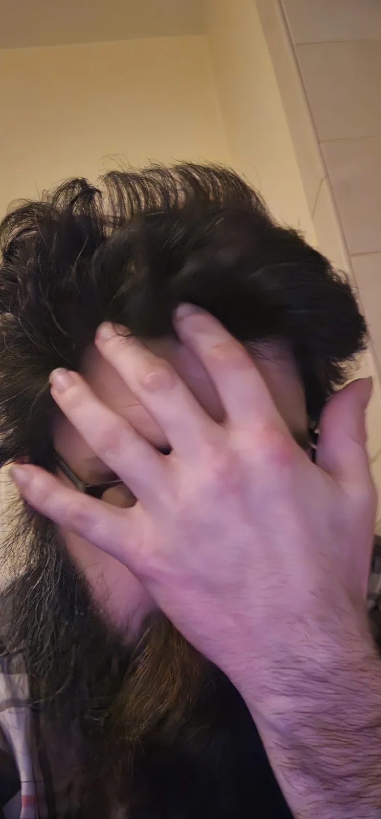 A person with long dark hair and glasses is shown from a close-up angle, covering their face with their right hand in a…