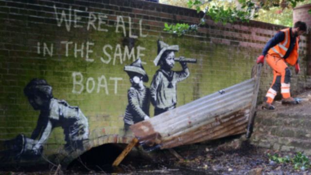 A mural depicting three children in a boat with the text 'WE'RE ALL IN THE SAME BOAT' is physically extended into reality by…