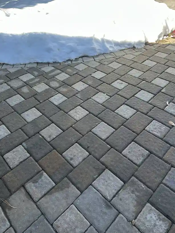 A close-up photo shows a herringbone-patterned stone patio with a thick, bright white snowbank melting along its edge…