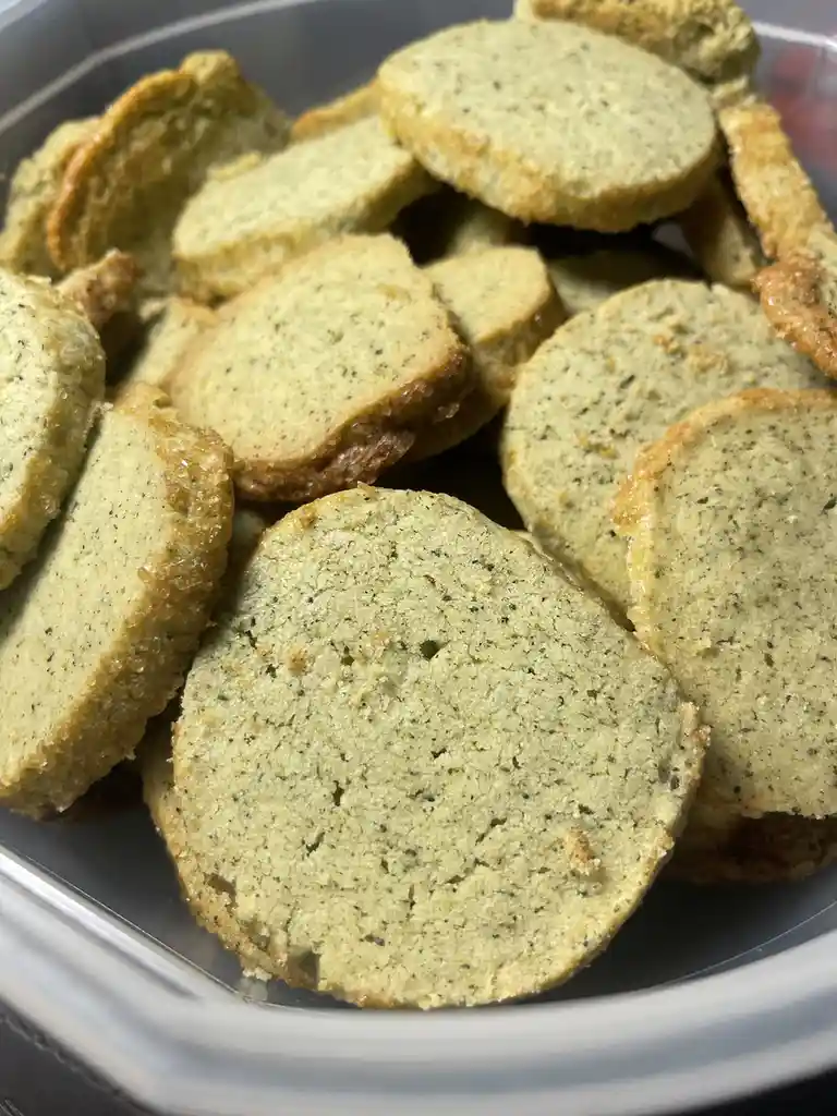 A close-up photo shows a pile of round, pale green, speckled crackers or biscuits in a clear plastic container. The text…