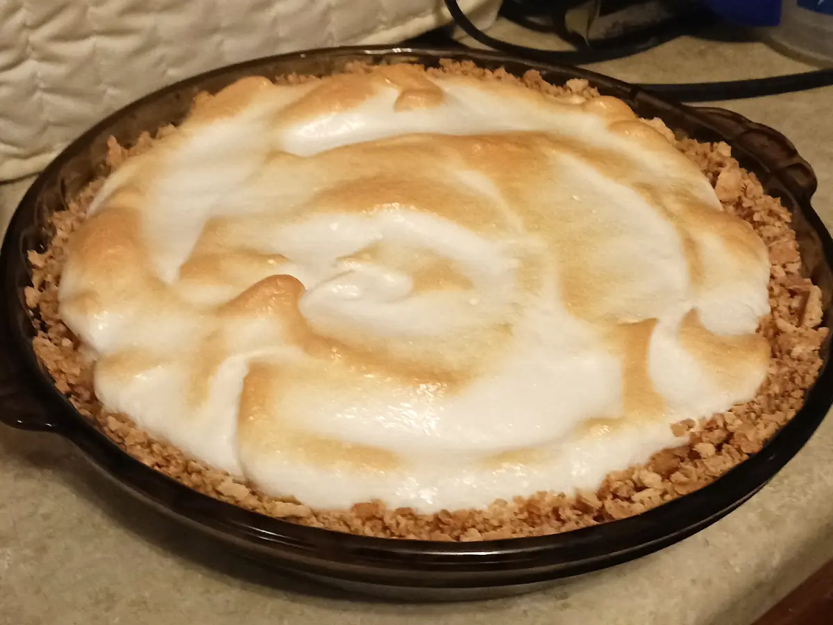 A close-up photo shows a baked dessert in a dark brown ceramic pie dish, featuring a golden-brown toasted meringue topping…