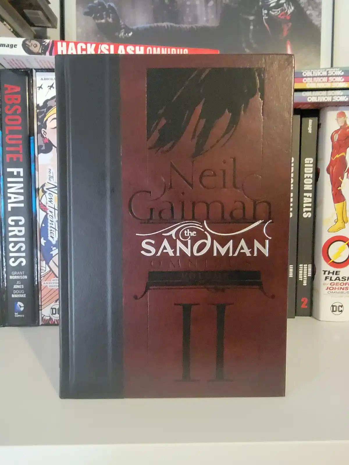 A close-up of a dark red and black hardcover comic book titled 'Neil Gaiman the Sandman Omnibus Volume II' standing upright…