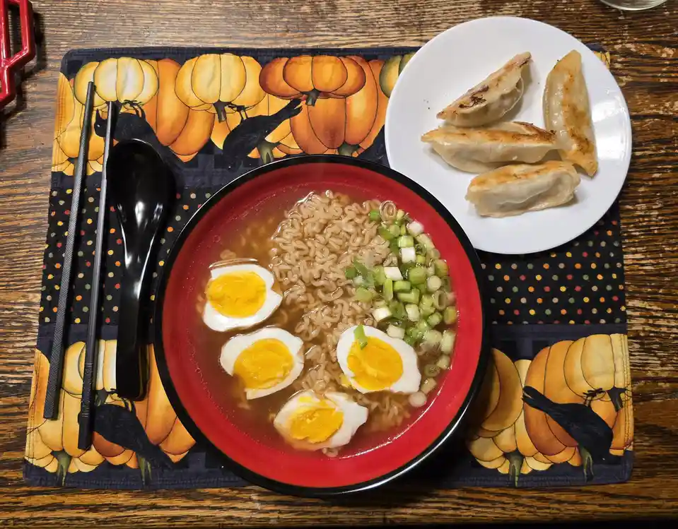 A top-down photo of a meal featuring a red bowl of ramen with four halved soft-boiled eggs and chopped green onions…
