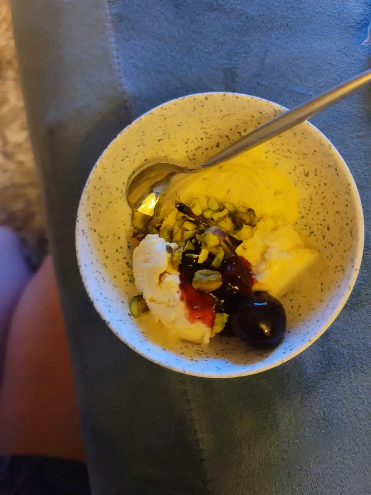 A top-down photo of a speckled ceramic bowl containing scoops of yellow and white ice cream, topped with chopped pistachios…