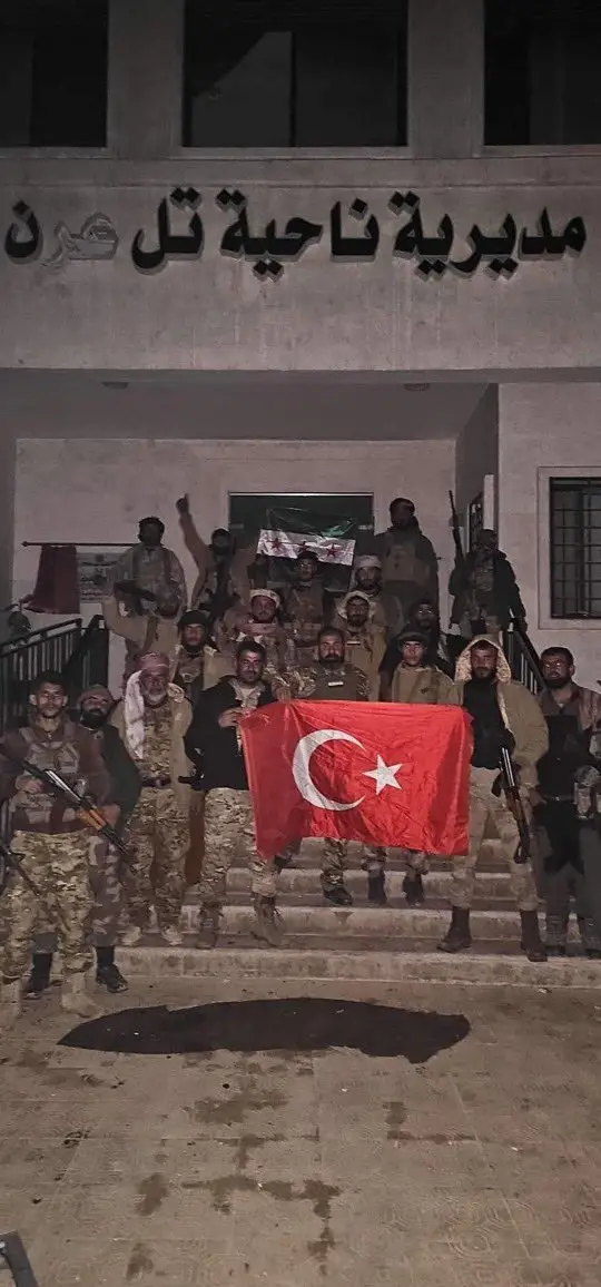 A group of armed men in camouflage uniforms stand on the steps of a building at night, holding a large Turkish flag. Above…