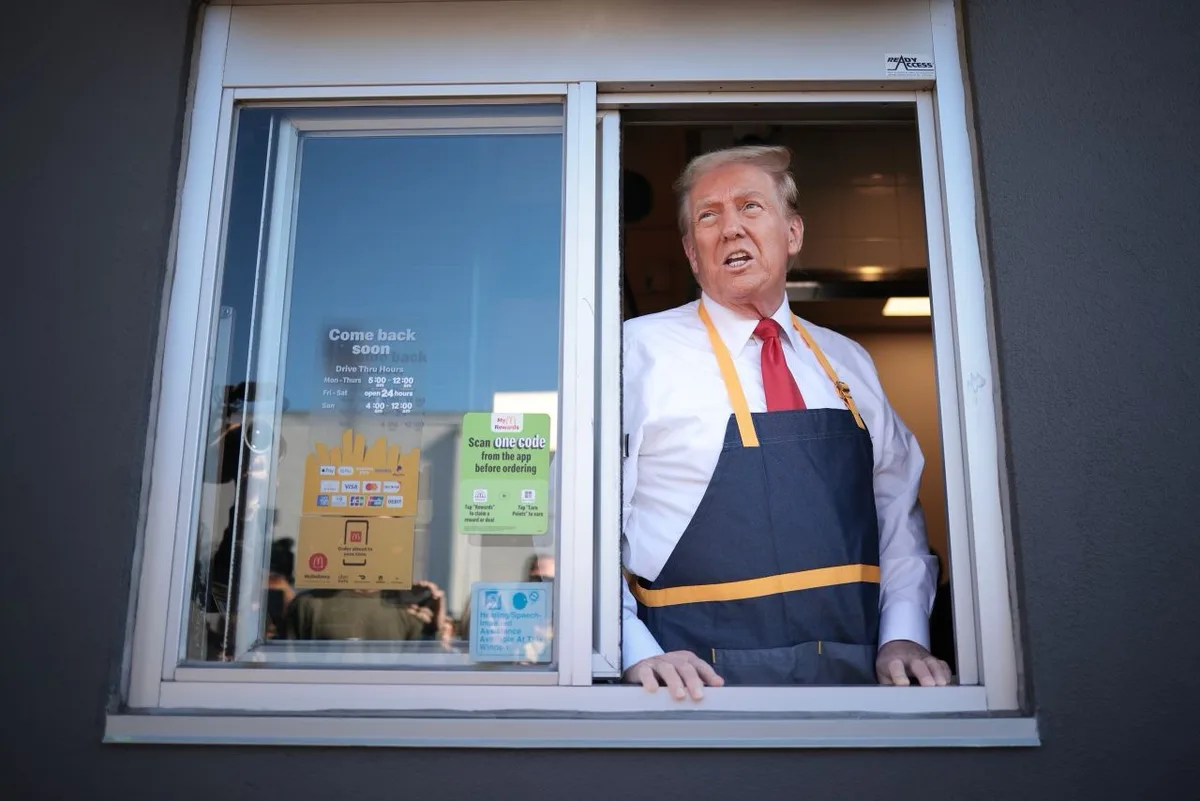A man with blond hair wearing a white shirt, red tie, and a blue and yellow McDonald's-style apron leans out of a drive-thru…