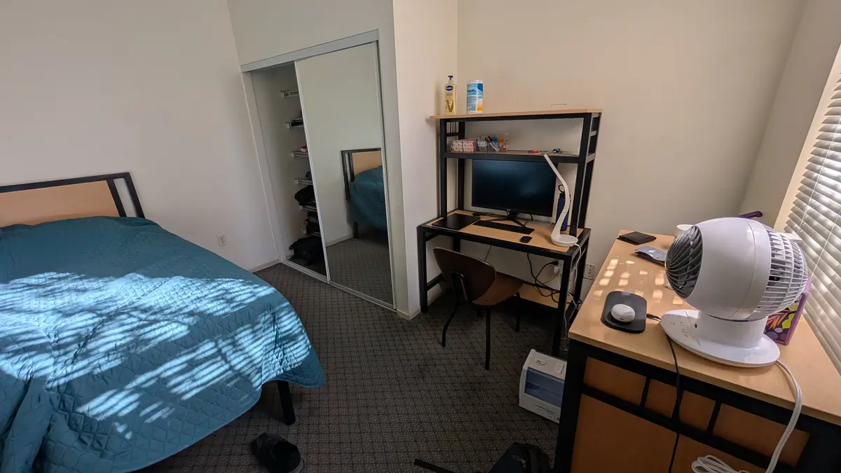 A tidy dorm room with a bed covered in a blue quilt, a desk with a computer and lamp, and a white oscillating fan on a…