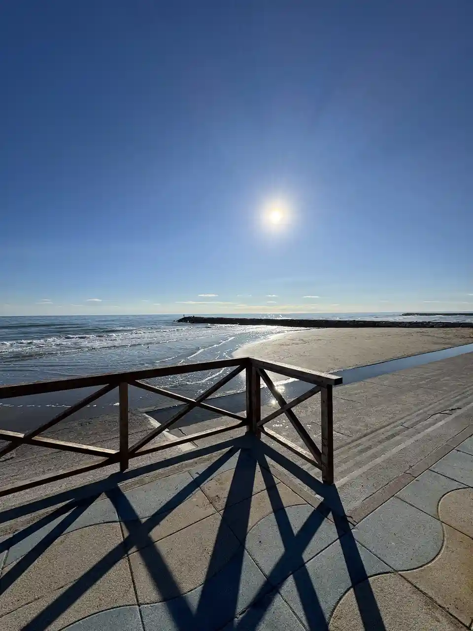 A bright, sunny beach scene with a wooden railing casting a sharp, geometric shadow on a tiled walkway. The sun is high in a…