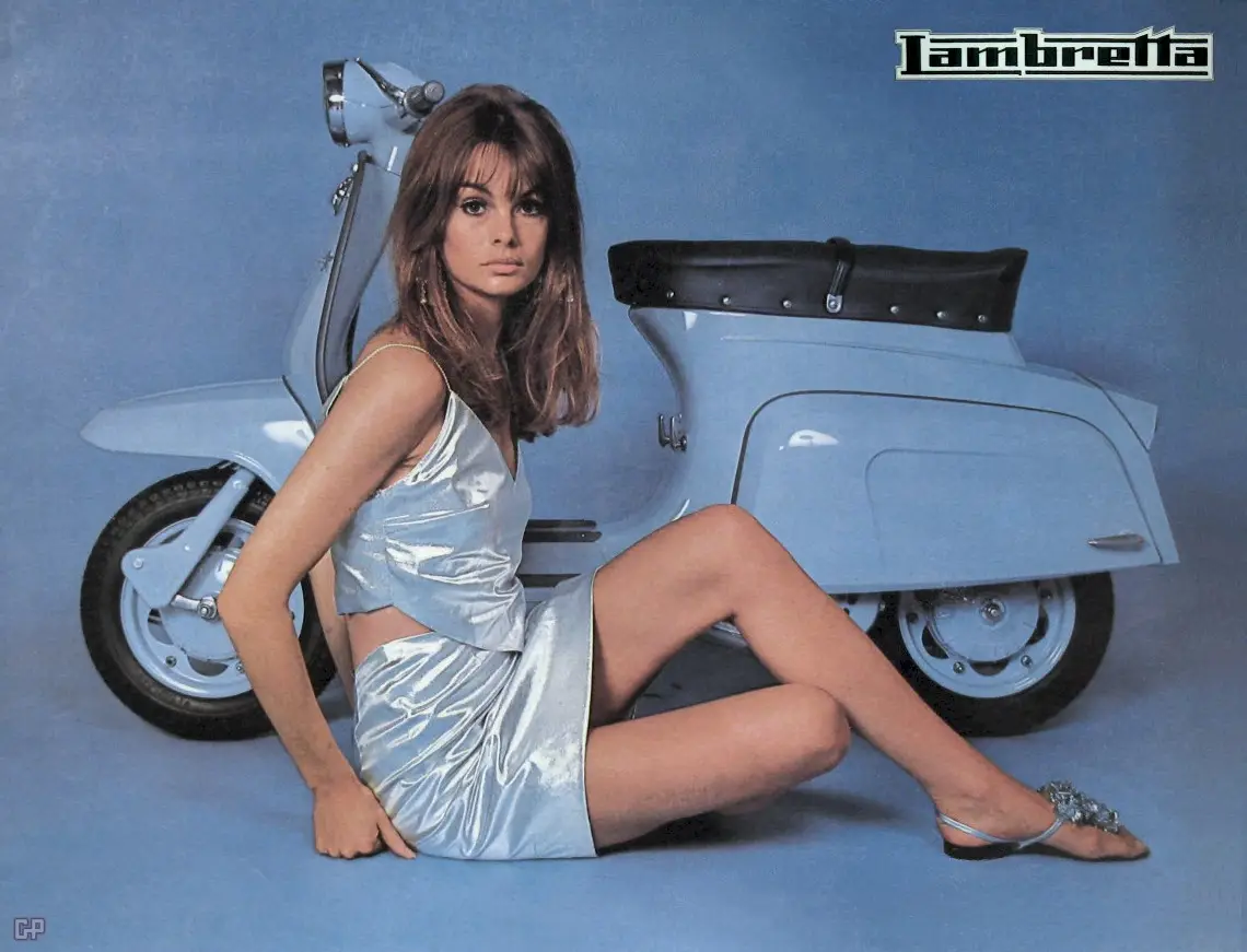 A vintage promotional photo shows a woman in a shiny silver dress sitting on a light blue Lambretta scooter against a solid…