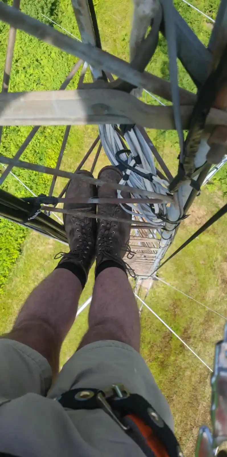 A first-person view from the top of a tall metal tower, showing a person's legs and brown work boots standing on a metal…