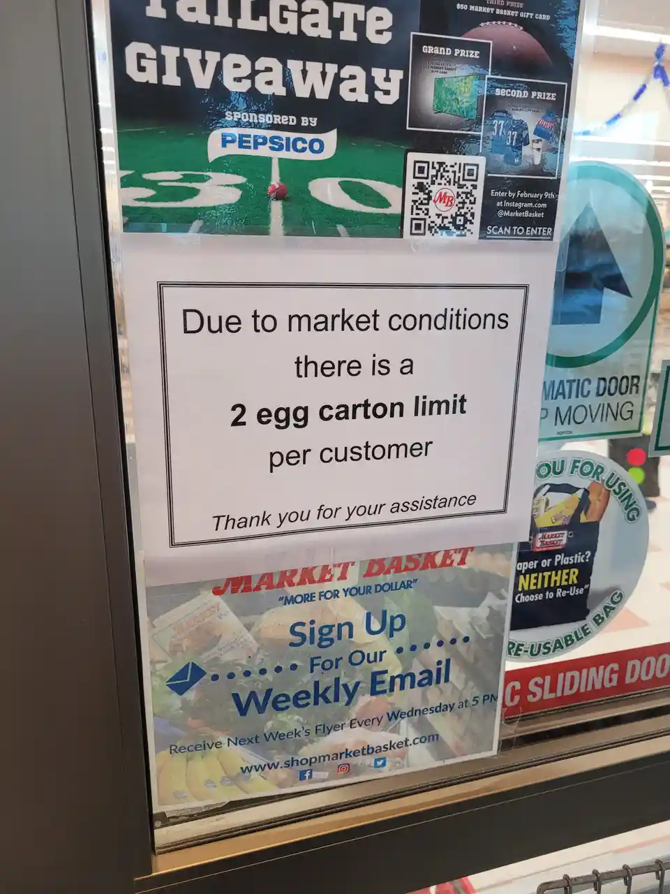 A printed sign taped to a store window states a '2 egg carton limit per customer' due to 'market conditions,' with a polite…