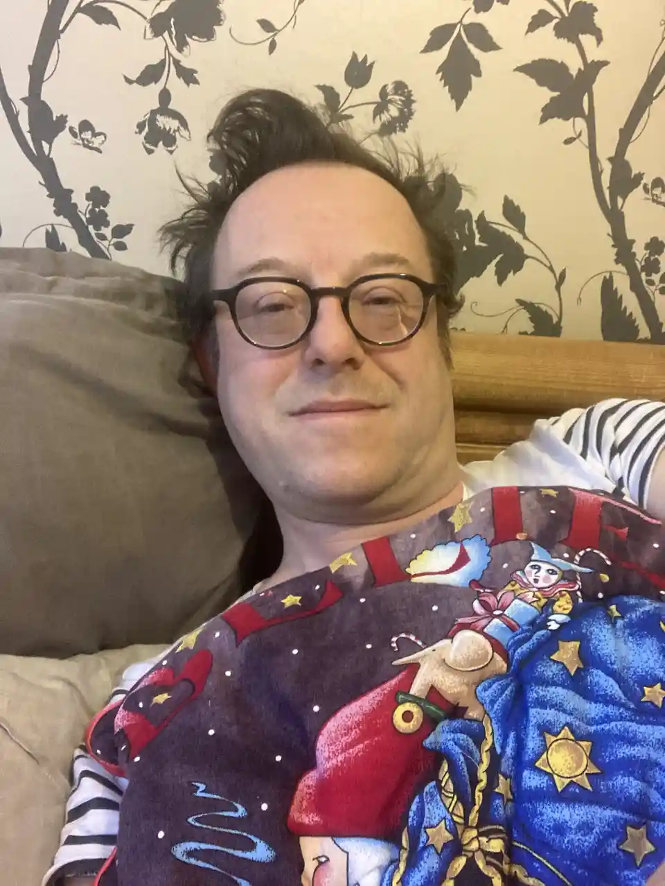 A man with glasses lies in bed under a colorful blanket featuring cartoon characters and stars, looking relaxed and slightly…