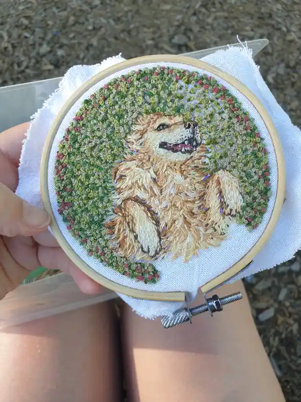 A hand holds a wooden embroidery hoop displaying a detailed, colorful embroidery of a fluffy golden dog with its mouth open…