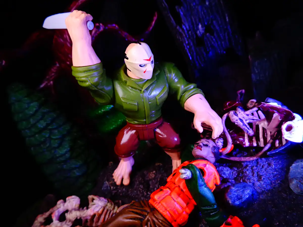 A toy action figure wearing a white mask with red markings and a green shirt stands over a fallen figure in an orange vest…