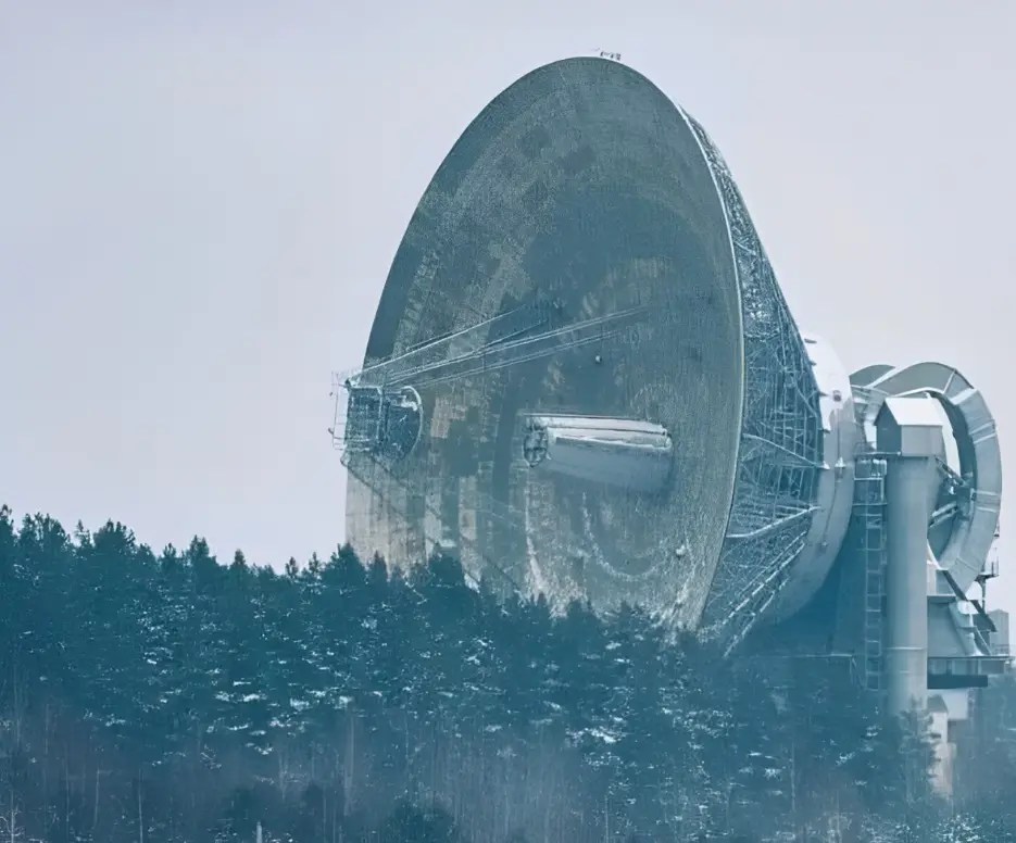 A large, dome-shaped radio telescope sits in a snowy forest, its metallic surface and complex support structure visible…