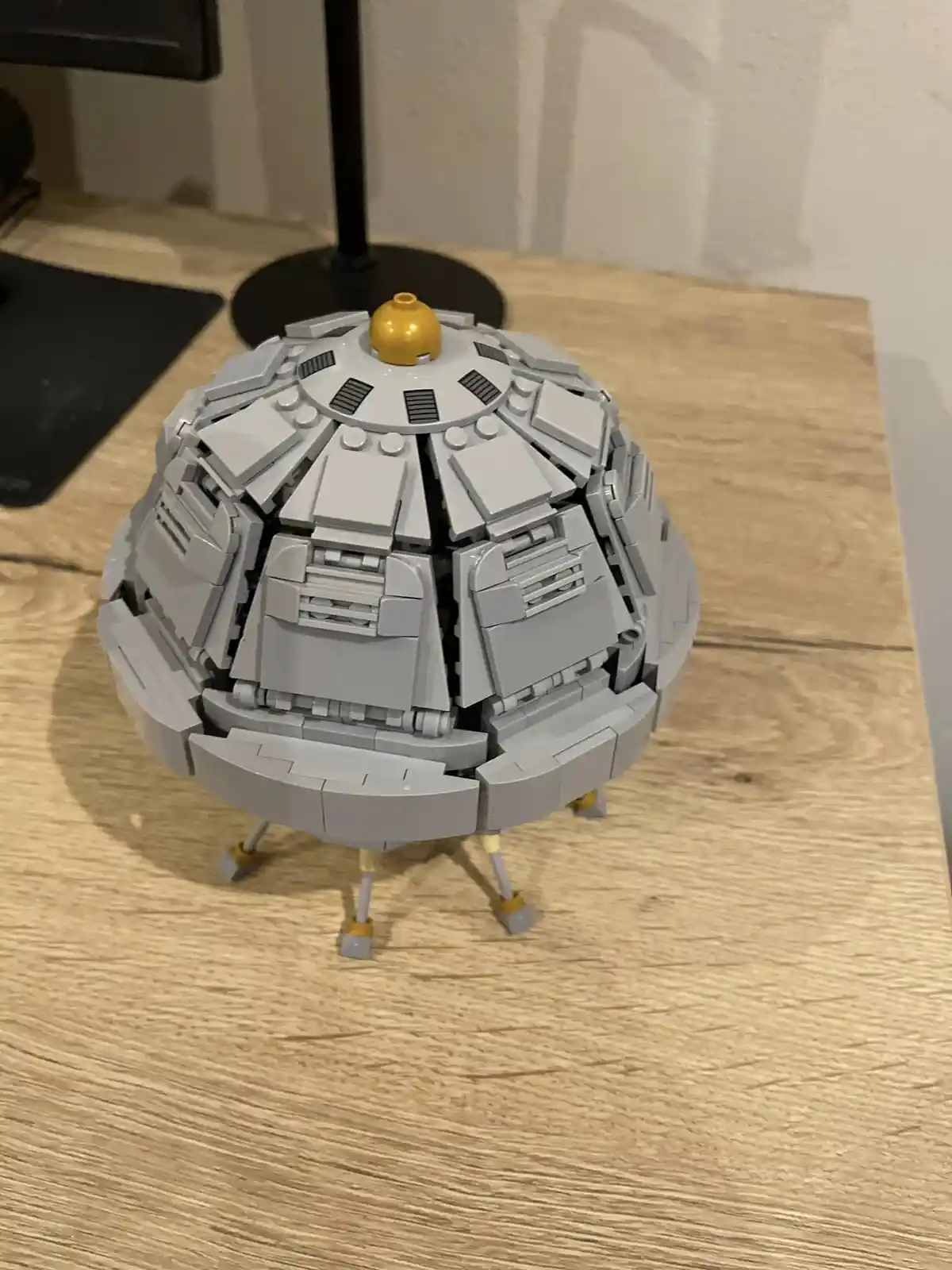 A grey Lego model of a spherical, dome-like structure with a yellow top piece, sitting on three legs on a wooden desk. The…