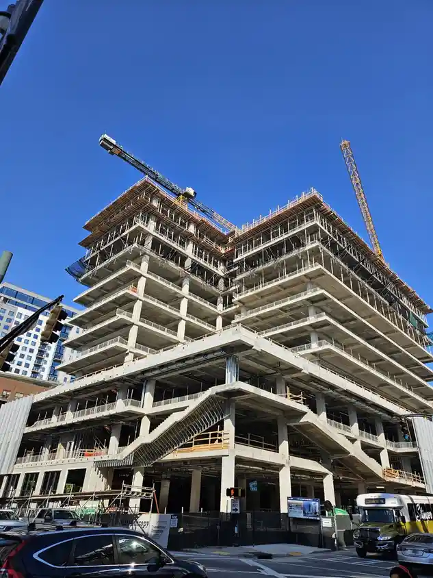 a low-angle photo of a tall, multi-story concrete building under construction against a clear blue sky, with cranes visible…