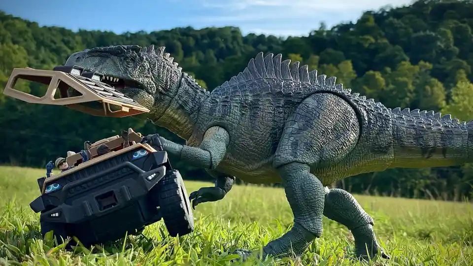 A large, grey toy dinosaur with a spiked back is shown outdoors in a grassy field, holding a toy armored vehicle in its…