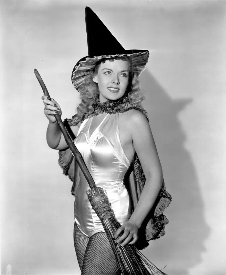 A vintage black-and-white photo shows a smiling woman in a witch costume with a pointed hat, holding a broomstick. She wears…