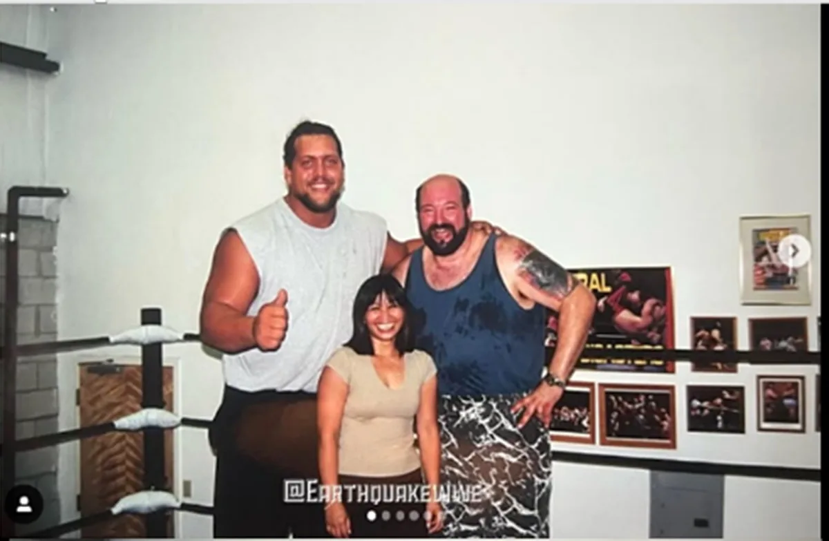 Three people pose together inside a wrestling ring, two large men flanking a smiling woman. The man on the left gives a…