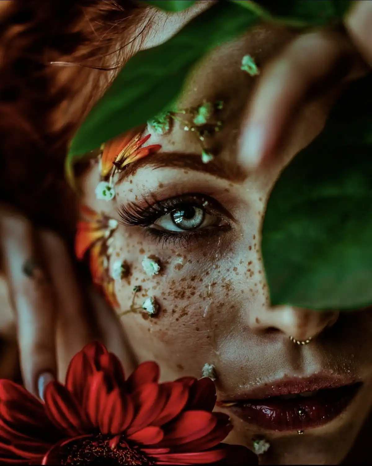 A close-up of a woman's face partially obscured by large green leaves and small white flowers, with freckles, a red flower…