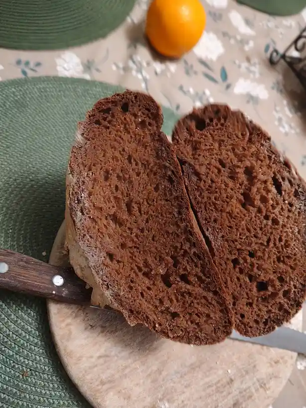 A close-up photo shows two thick slices of dark, porous rye or whole grain bread cut from a loaf, resting on a wooden…