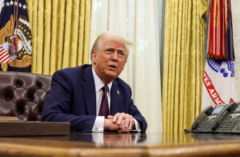 a man with white hair wearing a dark suit and tie sits at a large wooden desk in an office with gold curtains and…