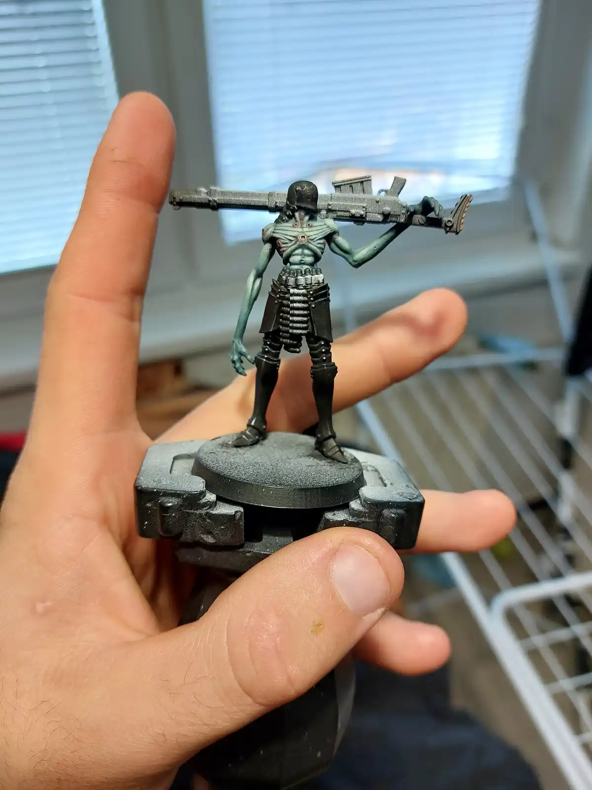 A hand holds a painted miniature figure of a grotesque, green-skinned humanoid with a helmet and a large, heavy weapon slung…