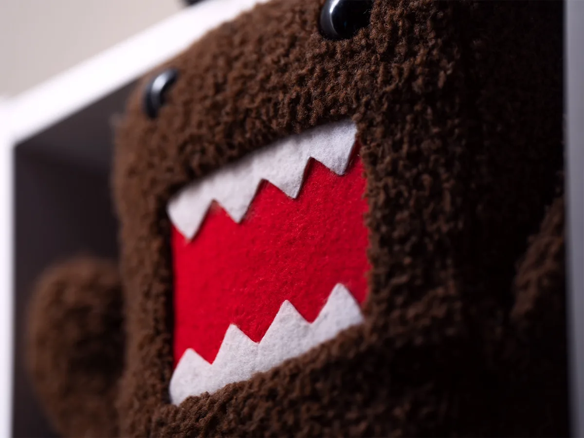 A close-up photo of a brown, fuzzy plush toy resembling Domo-kun, showing its wide, red mouth with white teeth. The overlaid…
