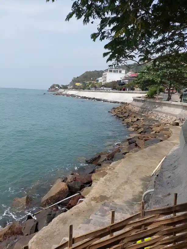 A scenic coastal view shows a calm sea next to a rocky shoreline with concrete seawalls and a wooden railing in the…