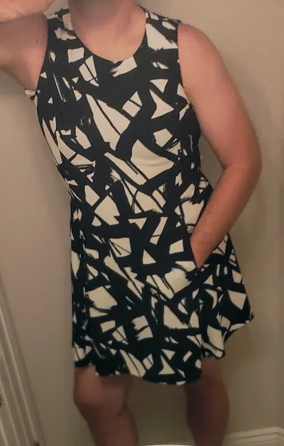 A person stands in front of a mirror wearing a black and white abstract-patterned sleeveless dress, one hand in their…