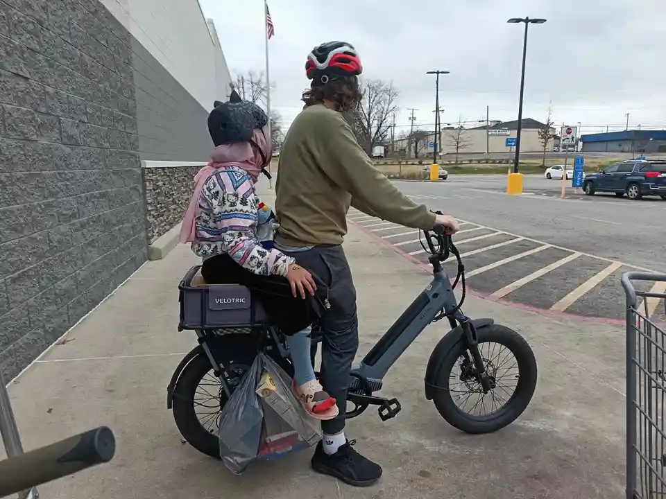 A person with long hair and a red helmet stands on a grey electric bike with a child seated in the rear cargo box. The child…