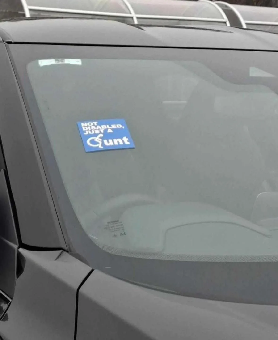 A close-up photo shows a blue sticker on the rear window of a gray car. The sticker reads 'NOT DISABLED, JUST A Cunt' in…