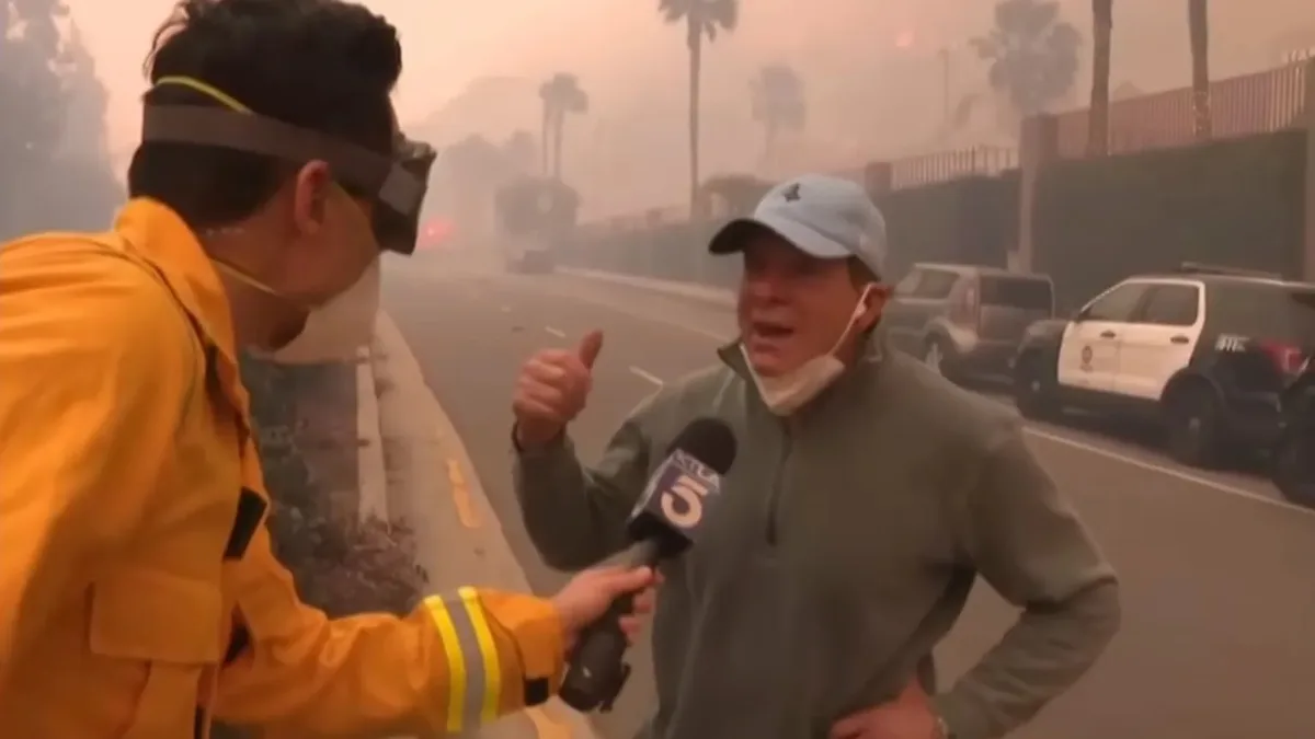 A man in a grey jacket and light blue cap, with a face mask pulled down, gives a thumbs-up while being interviewed by a…