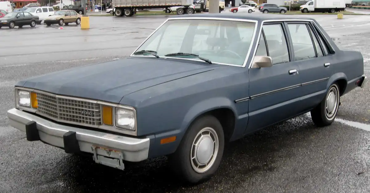 A blue 1980s Ford sedan is parked in a wet parking lot, looking dusty and slightly dented. The overlaid text reads 'i'm not…