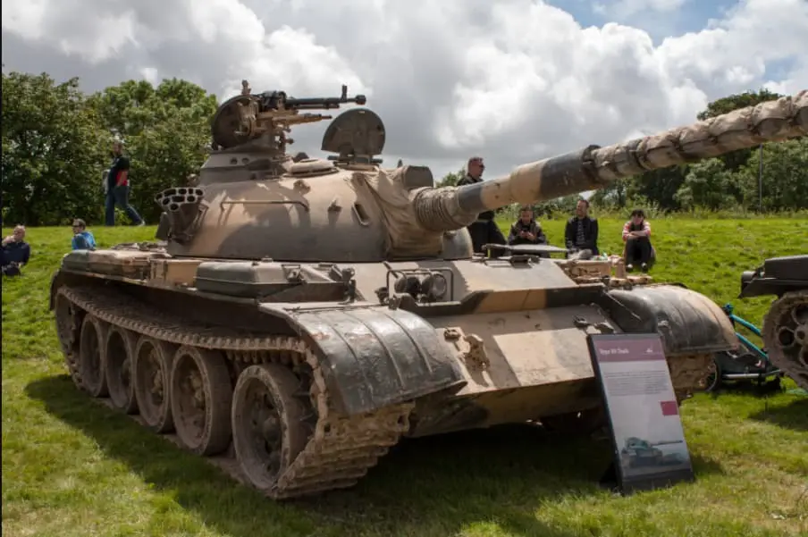 A large, dusty Type 59 tank is displayed outdoors on grass under a cloudy sky, with several people sitting and standing…