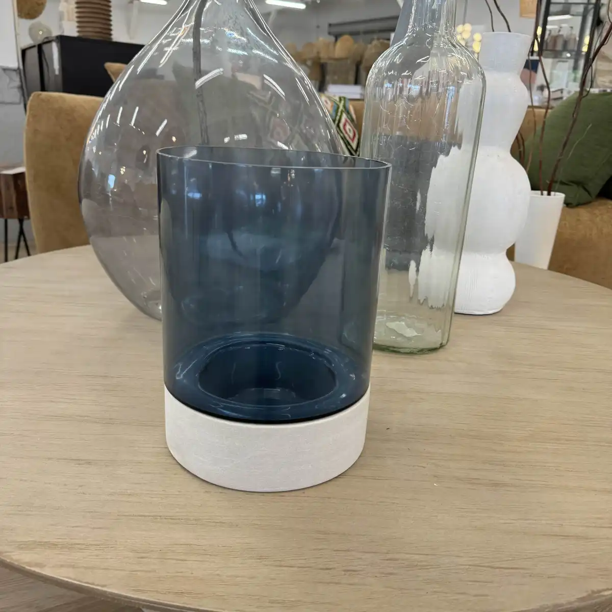 A dark blue glass vase with a white textured base sits on a light wooden table, surrounded by other decorative glass and…