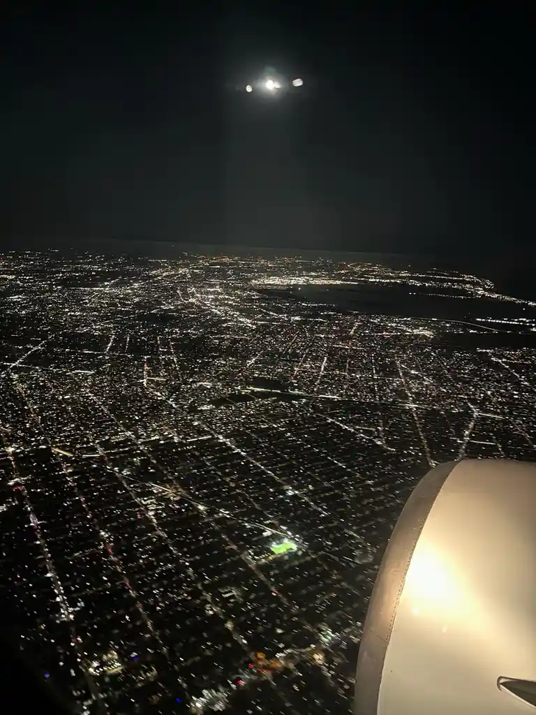 A nighttime photo taken from an airplane window shows a sprawling cityscape below illuminated by countless lights, with the…