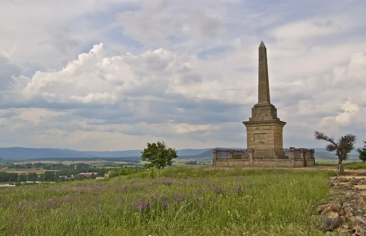 A tall stone obelisk monument stands alone on a grassy hilltop under a cloudy sky, surrounded by wildflowers and distant…