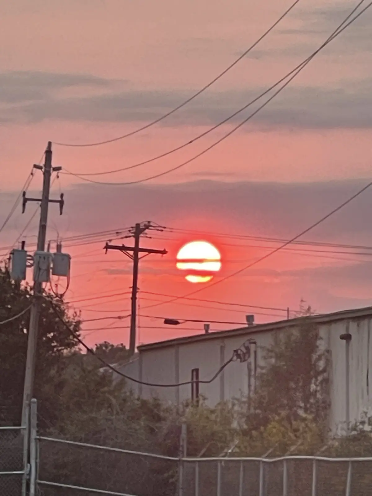 A sunset photo shows a red-orange sun partially obscured by clouds, with utility poles and wires crossing the frame. The sun…