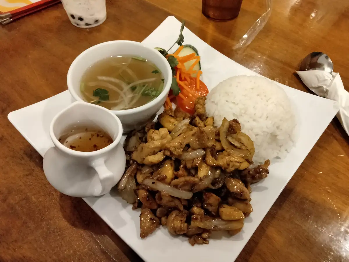 A plate of Vietnamese-style food featuring stir-fried chicken with onions, a mound of white rice, a bowl of clear broth soup…