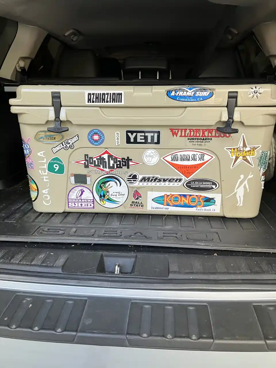 A beige Yeti cooler sits in the trunk of a Subaru, covered in numerous surfboard shop stickers like 'South Coast…