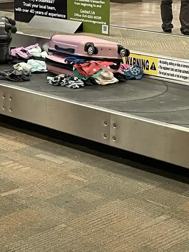 A pink suitcase sits open on an airport baggage carousel, its contents—various colorful clothes—spilled out and scattered…