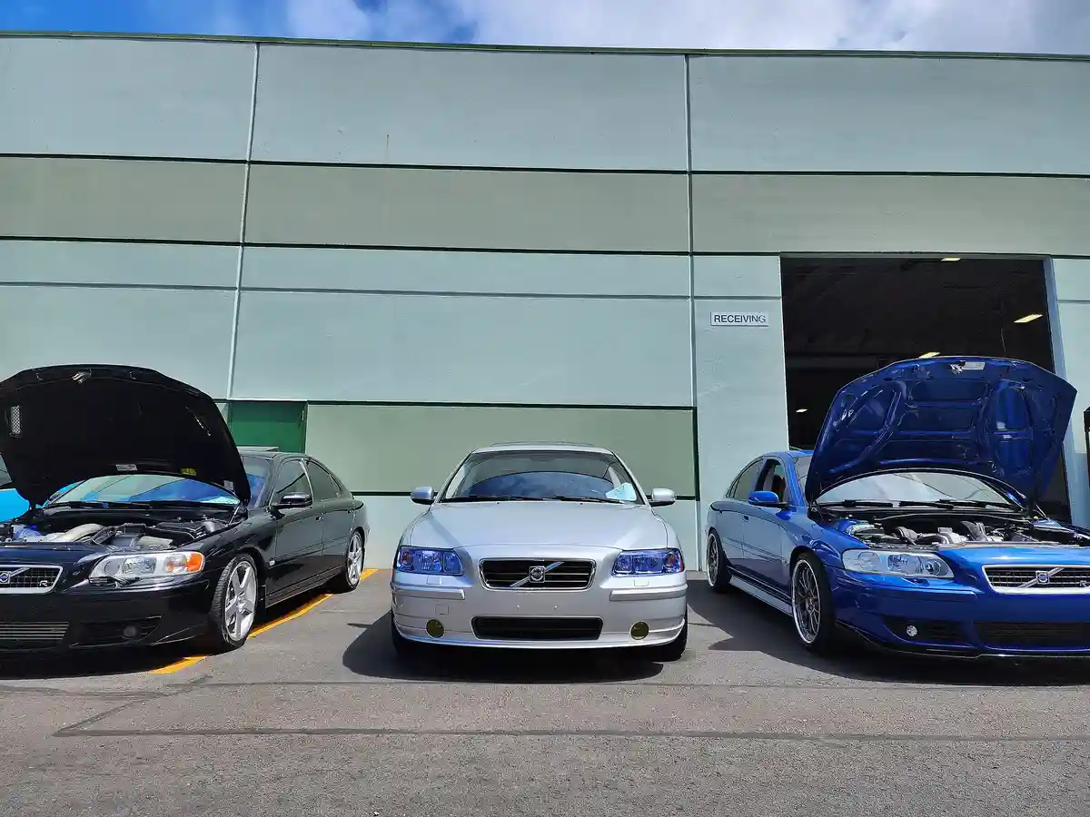 Three Volvo S80 R sedans are parked in front of a warehouse with a 'RECEIVING' sign; the black and blue cars on the sides…
