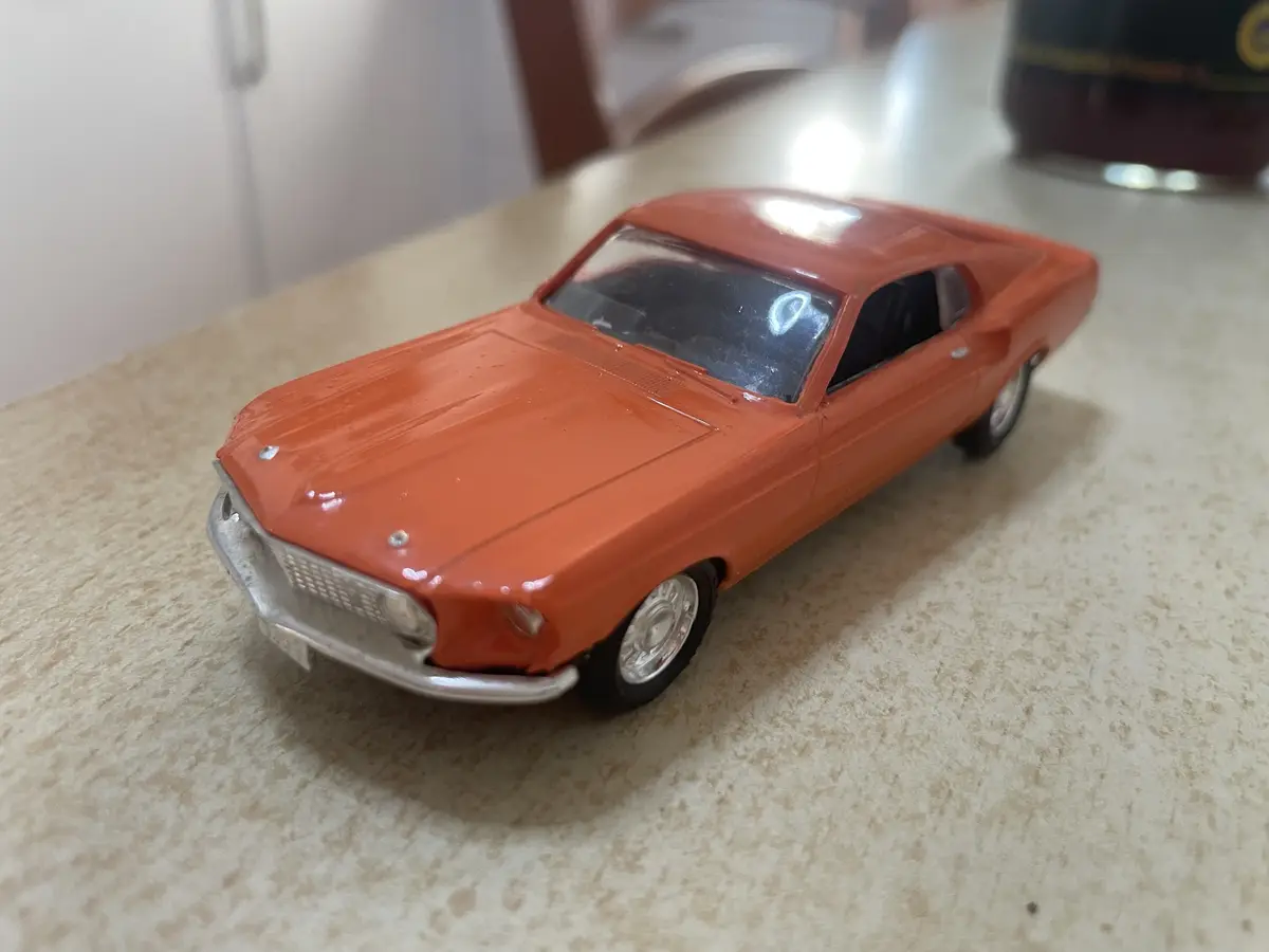 A small orange toy car with a silver grille and chrome wheels sits on a speckled countertop. The car has a classic muscle…