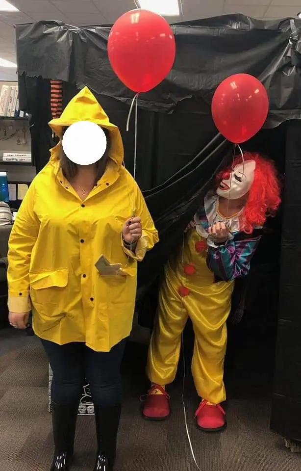 A person in a bright yellow raincoat and black boots stands beside a clown in a yellow suit and red wig, both holding red…