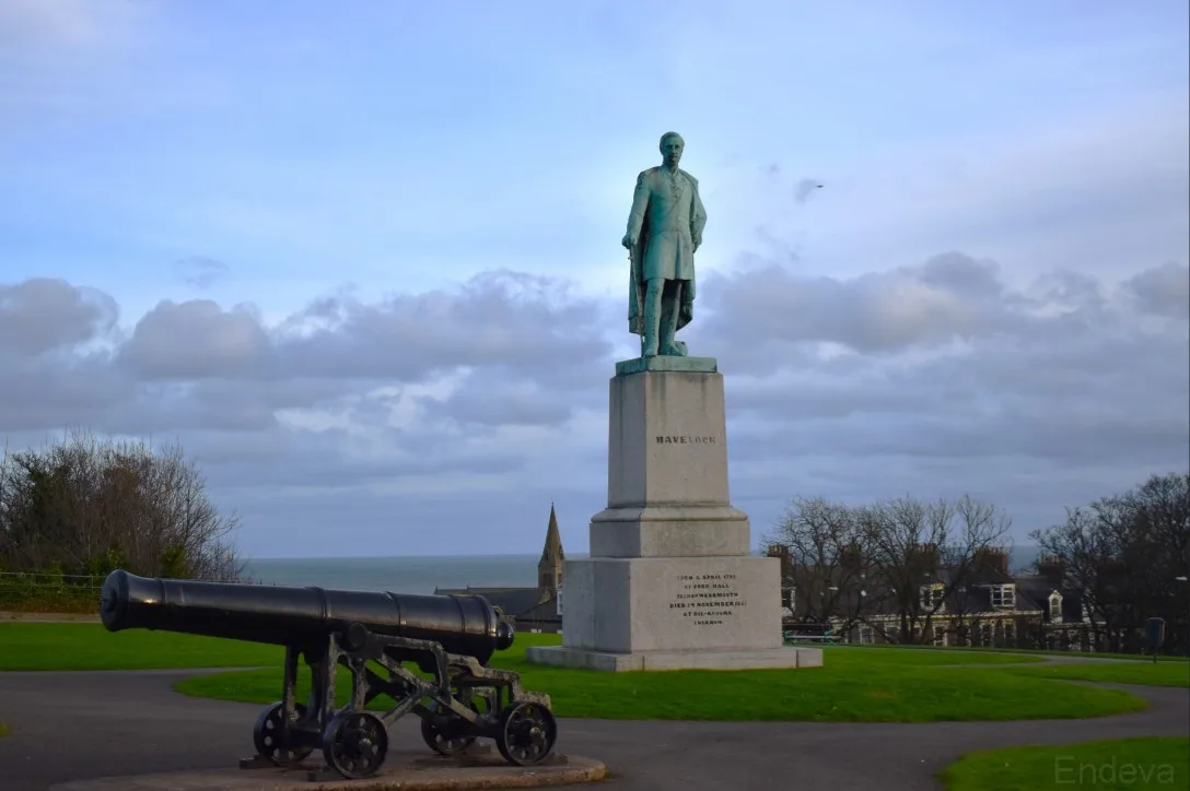 A bronze statue of a man named Havelock stands on a stone pedestal overlooking the sea, with an old black cannon positioned…