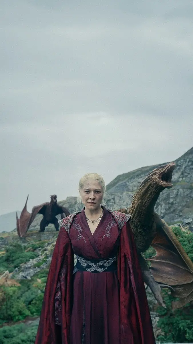 A woman with platinum blonde hair wearing a red robe stands in a mountainous, rocky landscape. Two large, brown dragons are…