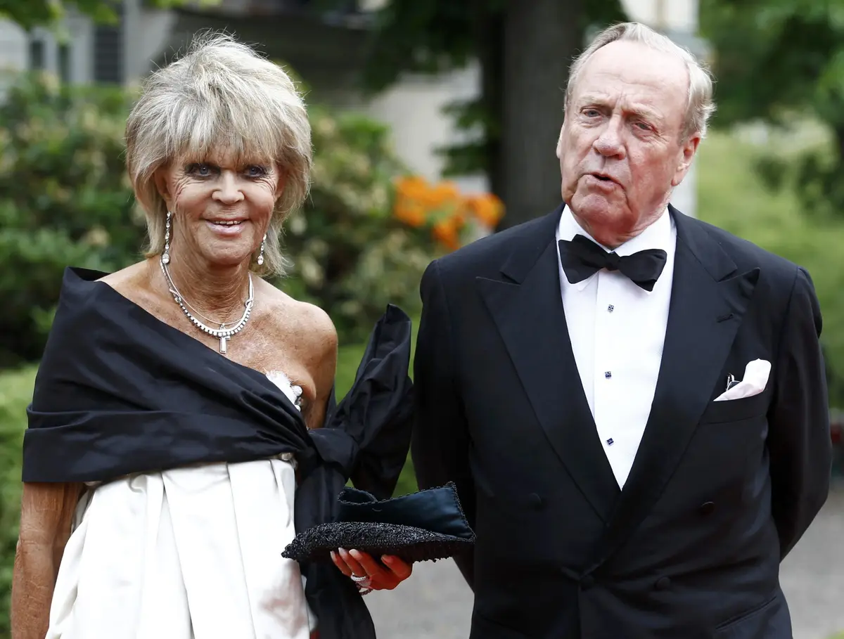 An older woman in a white dress with a black shawl smiles while holding a clutch, standing next to an older man in a tuxedo…
