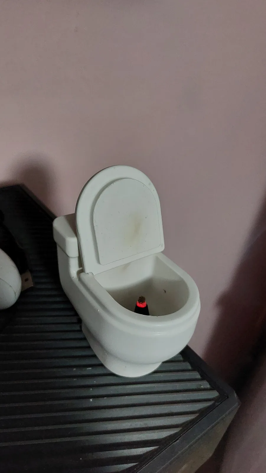 A small white toy toilet with its lid open sits on a black slatted surface against a pink wall. Inside the toilet bowl, a…